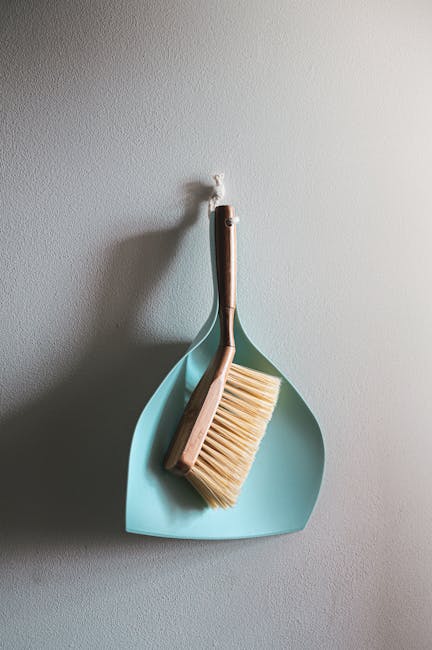 A cleaning brush with a wooden handle and natural bristles hangs on a wall hook, positioned inside a light blue dustpan. The brushes and dustpan are clean and well-maintained, with the brush angled ag