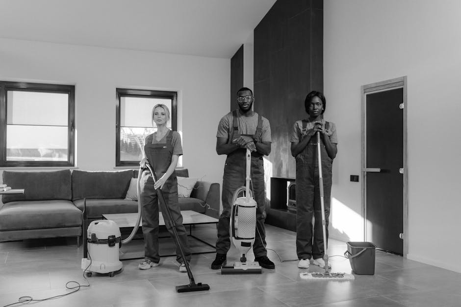 Three professional cleaners from Camden Cleaners are performing surface cleaning and deep cleaning in a modern living room. The room features a large, L-shaped fabric sofa positioned against a wall wi